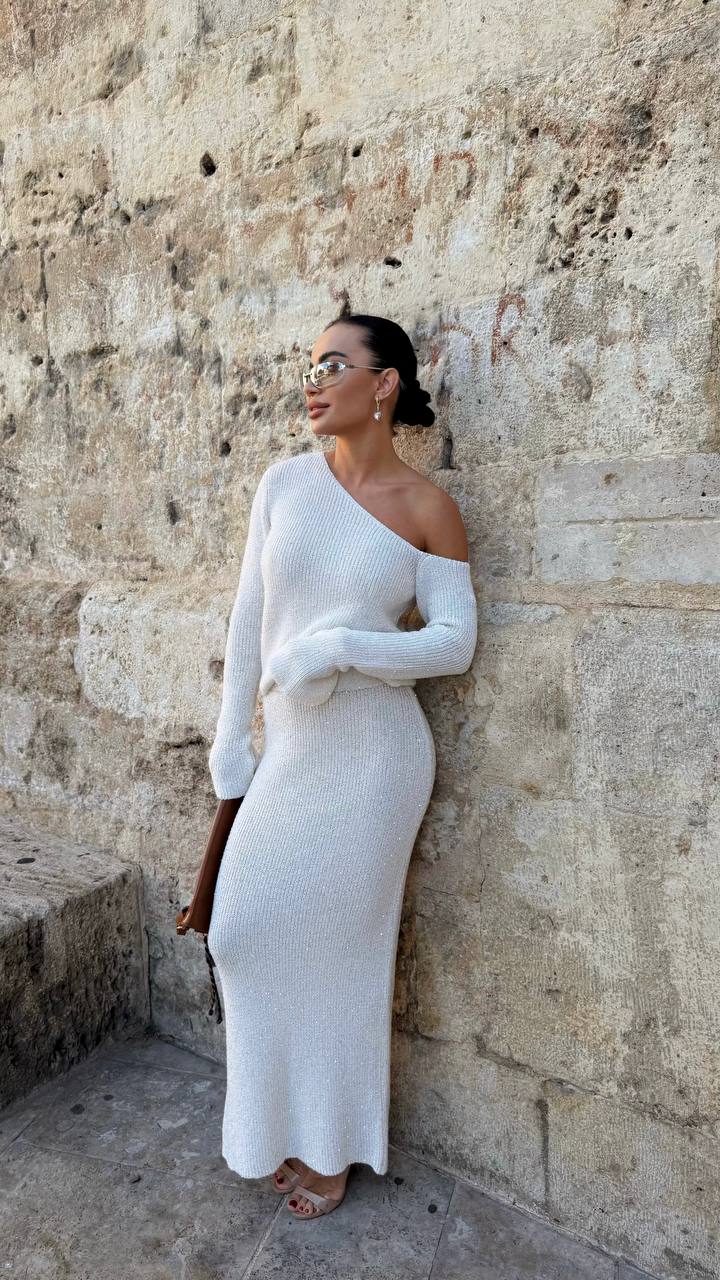 Woman in a white one-shoulder dress standing against a textured stone wall.