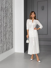 A woman models a white long-sleeve midi dress with a textured crochet pattern and button-front closure, standing in a modern gray hallway with a patterned wall panel on the left.