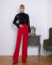 Person wearing a black top and red pants standing in a room with a side table and chair.