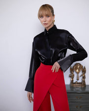 Woman wearing a black satin blouse and red pants against a white wall.