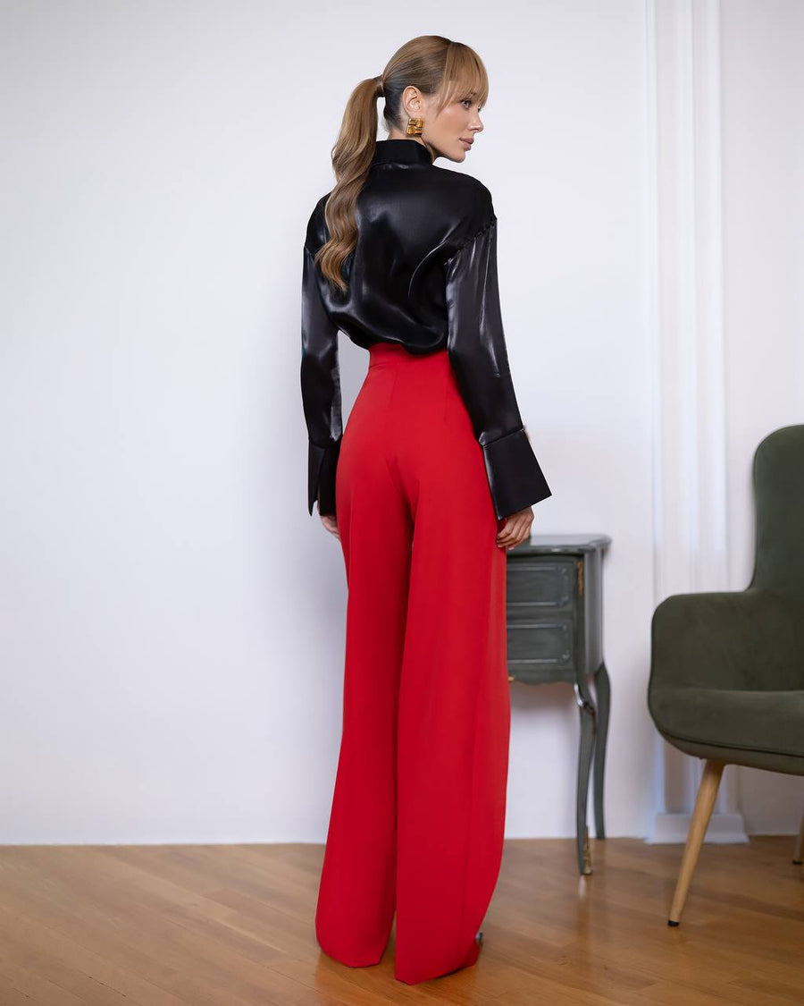 Person wearing a black top and red pants standing in a room with a chair and table.