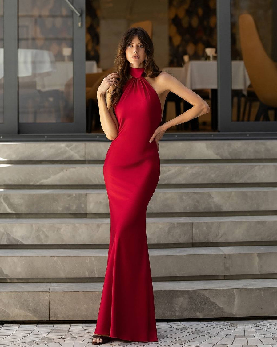 A woman modeling a sleeveless, high-neck, floor-length red gown standing on stone steps in front of a glass-front building.