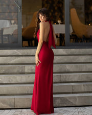 A woman wearing a floor-length red gown with a draped open back, standing on stone steps in front of a glass-front building.
