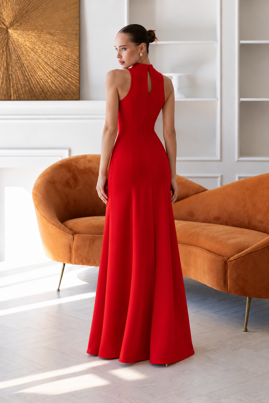 A woman wearing a bright red sleeveless floor-length gown stands with her back to the camera in a bright, modern room with an orange velvet sofa and white paneled walls.