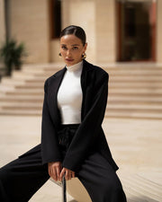 Woman in a black coat and white turtleneck sitting on steps outdoors.