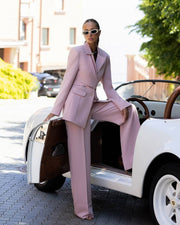 A woman wearing a light pink tailored suit and white sunglasses sits in the open door of a white convertible car on a paved street.