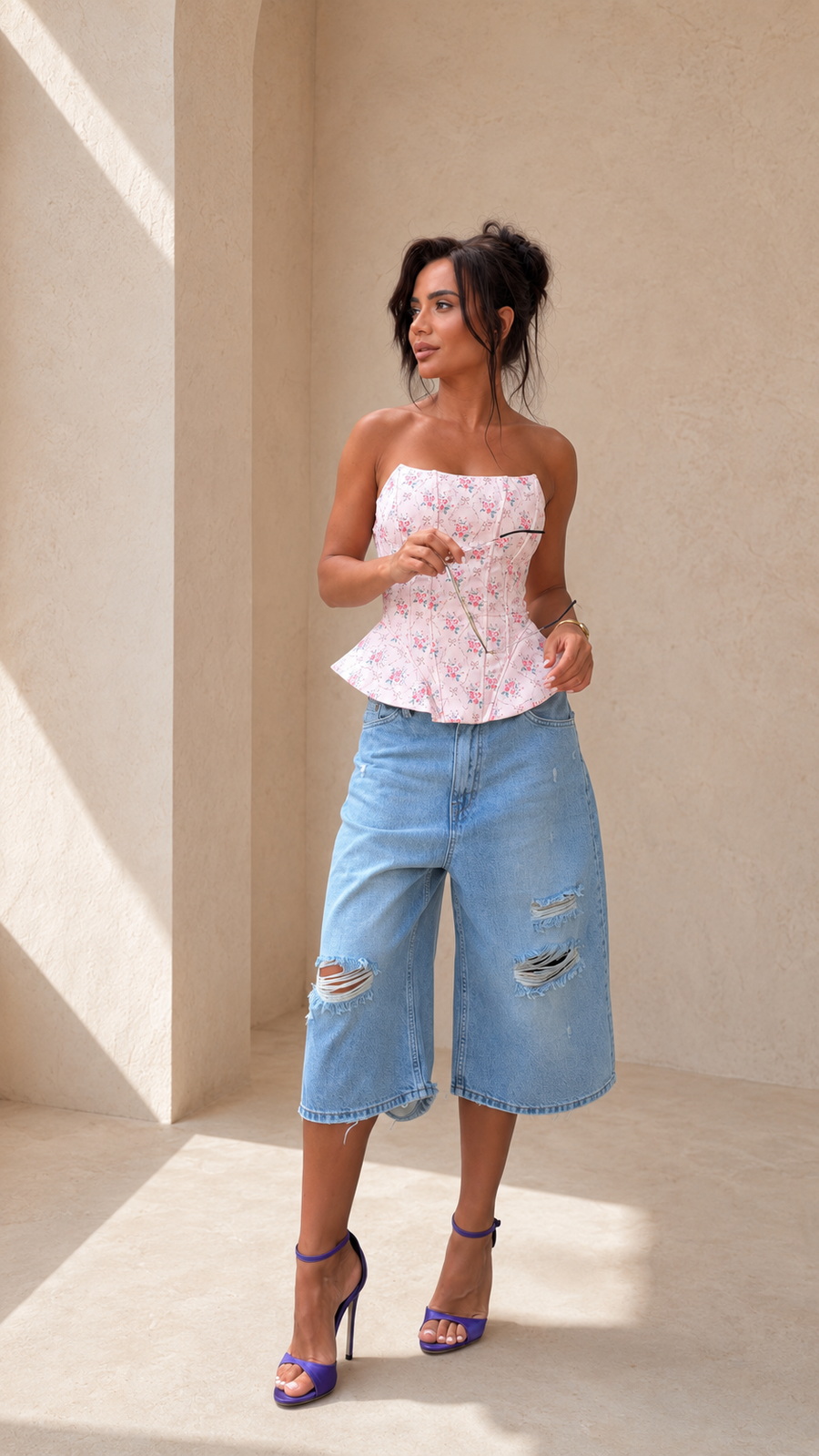 A woman modeling a strapless peplum top with a pink and white floral pattern, paired with light-wash distressed denim culottes and purple high heels, standing against a beige stucco wall.