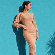 A woman wearing a fitted, long-sleeve mock-neck midi dress in a warm beige or camel suede-like fabric, posing against a bright turquoise textured wall.