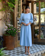 Woman in a light blue dress standing in front of a decorative entrance with plants.