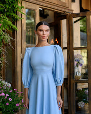 Woman in a light blue dress standing in front of a wooden door with plants around.