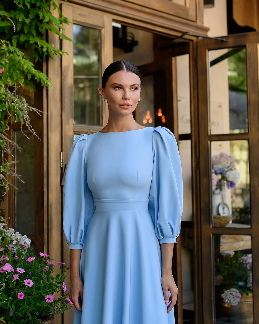 Woman in a light blue dress standing in front of a wooden door with plants around.