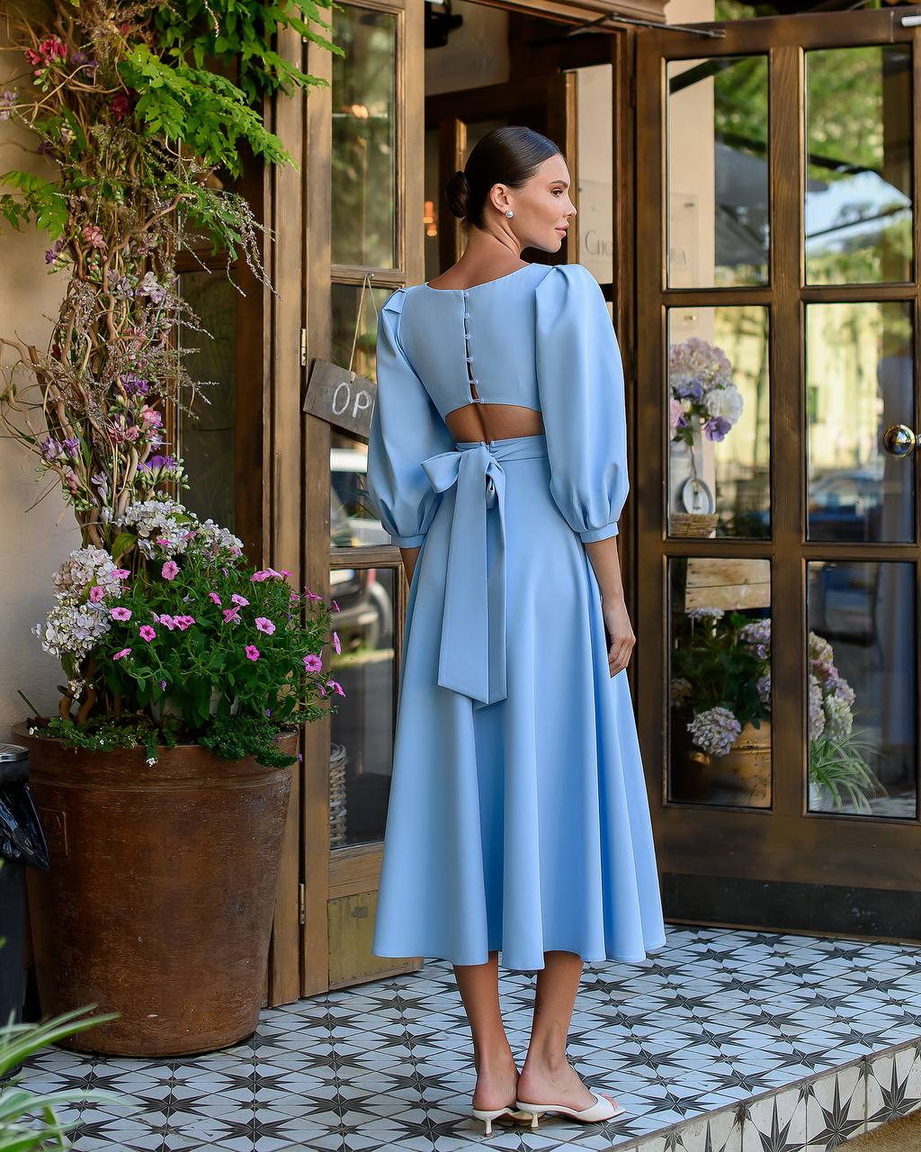 Woman in a light blue dress standing in front of a store entrance with flowers and plants around.