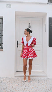Woman wearing a red floral dress with white accents standing in front of a white door.