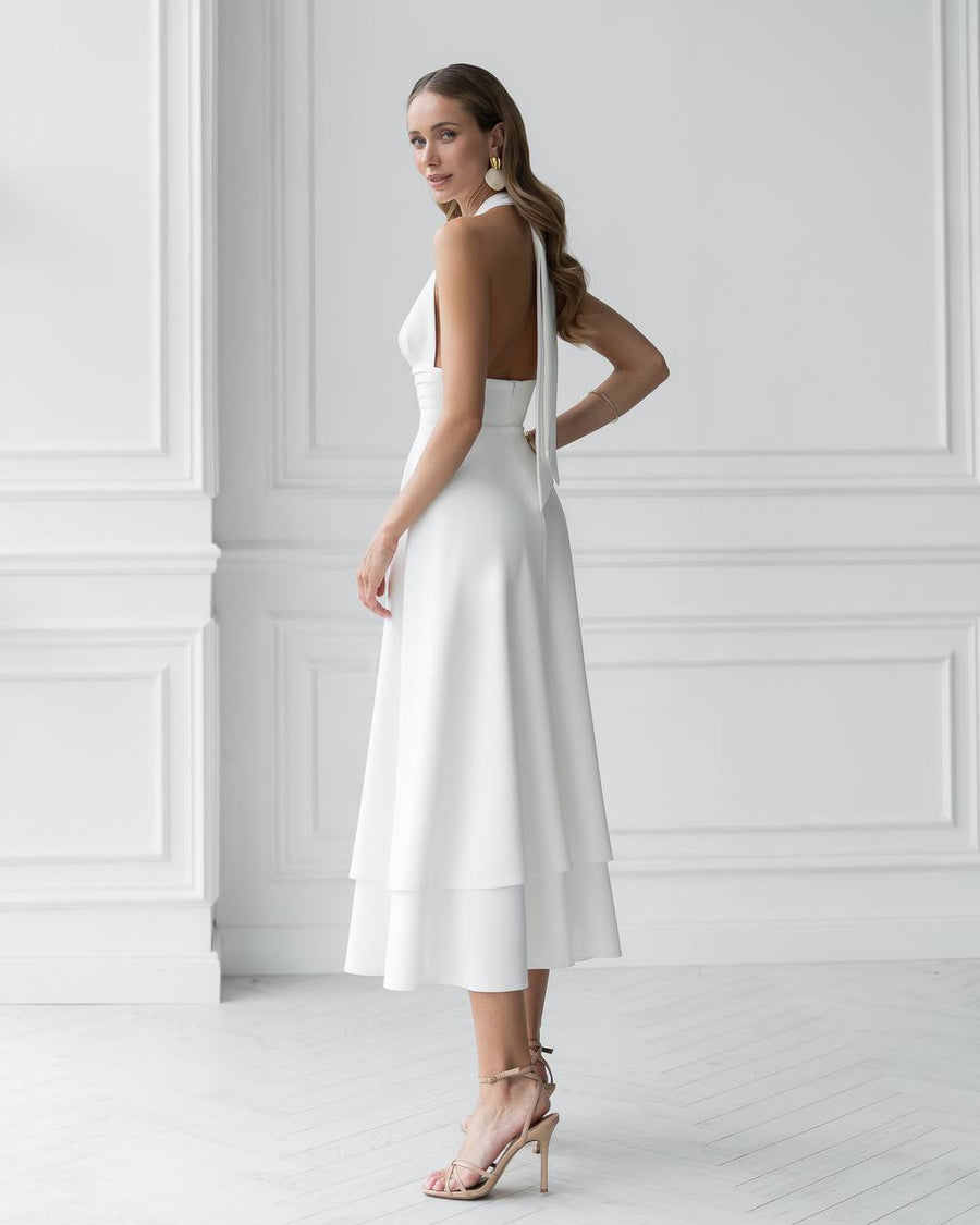 Woman wearing a white halter neck dress with thin straps against a white wall.