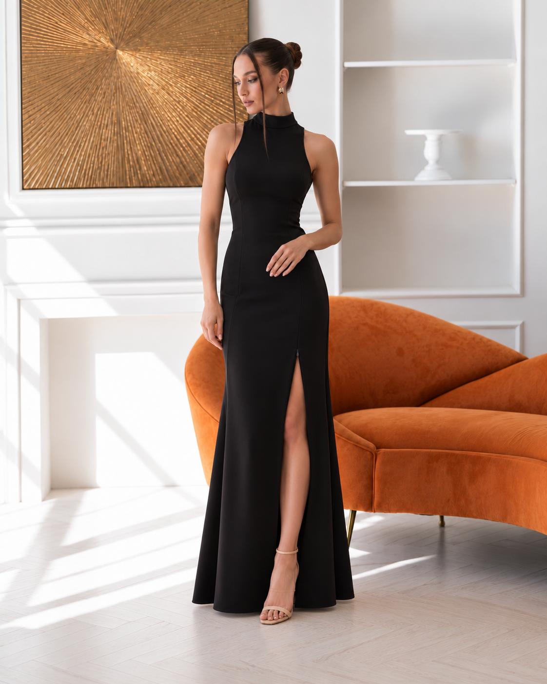 A woman models a sleeveless black high-neck floor-length gown with a thigh-high slit, standing in a bright room with a white wall, shelving, and an orange curved sofa.
