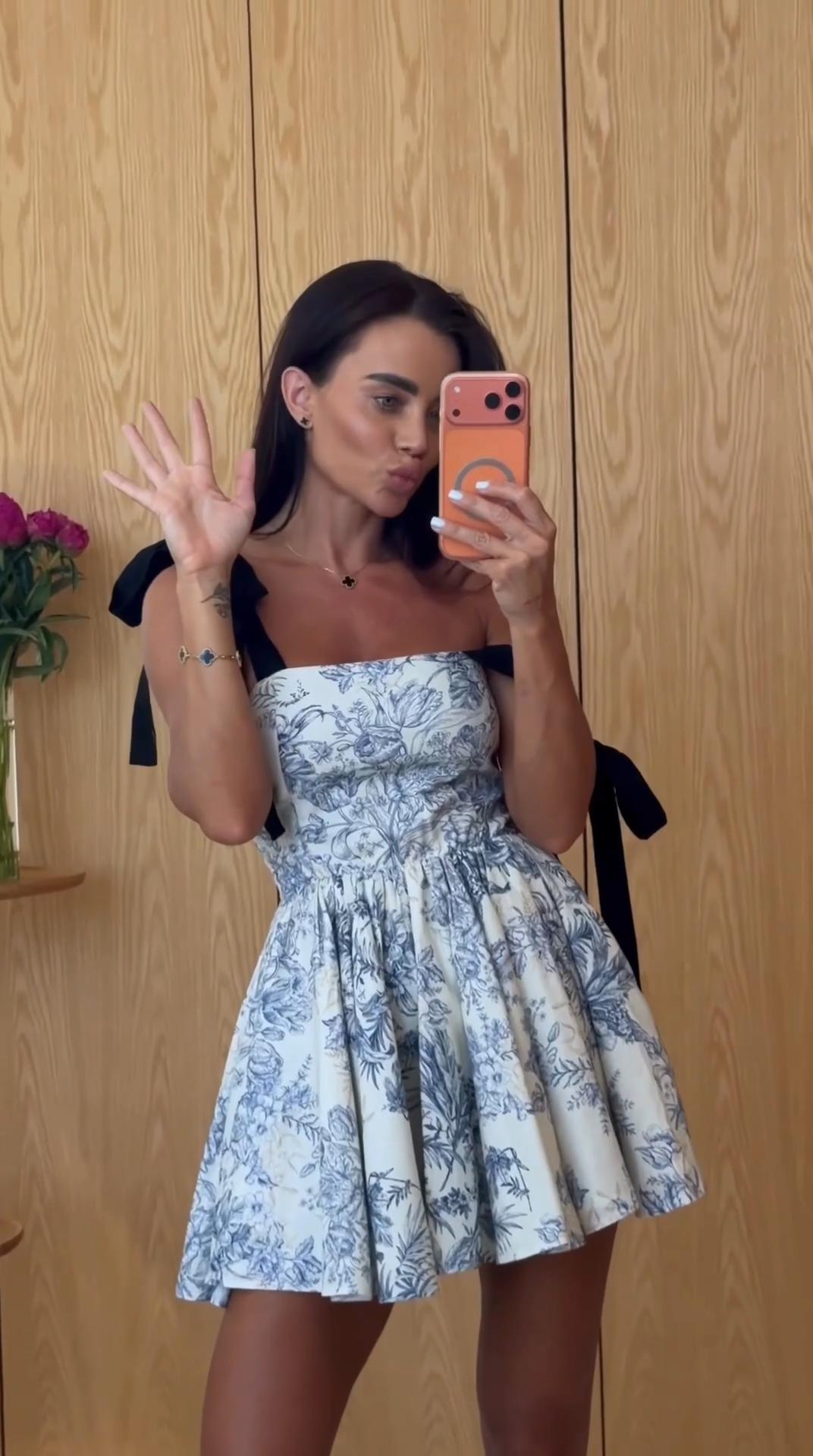 A woman takes a mirror selfie wearing a strapless white dress with a blue toile floral print and black shoulder ties, standing in front of a light wood-paneled wall.