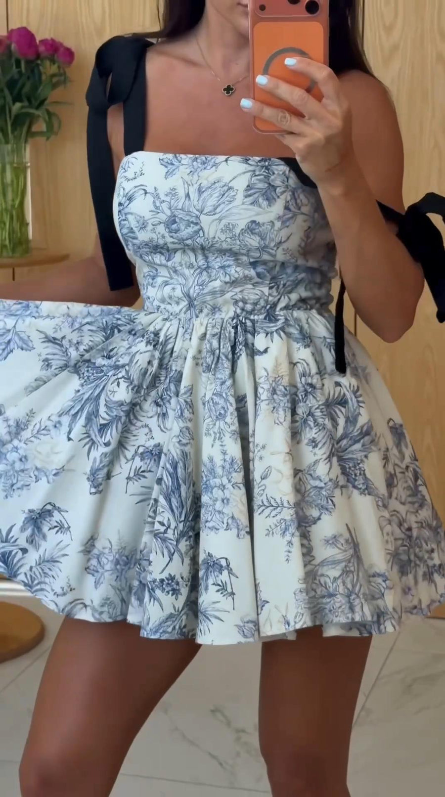 A woman takes a mirror selfie wearing a strapless white dress with a blue toile floral print and black shoulder ties, standing in an indoor room with a wooden door and flowers in the background.