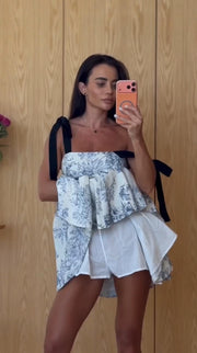 A woman takes a mirror selfie wearing a white ruffled top with a blue floral pattern and black shoulder ties, paired with white shorts, against a light wood-paneled wall.