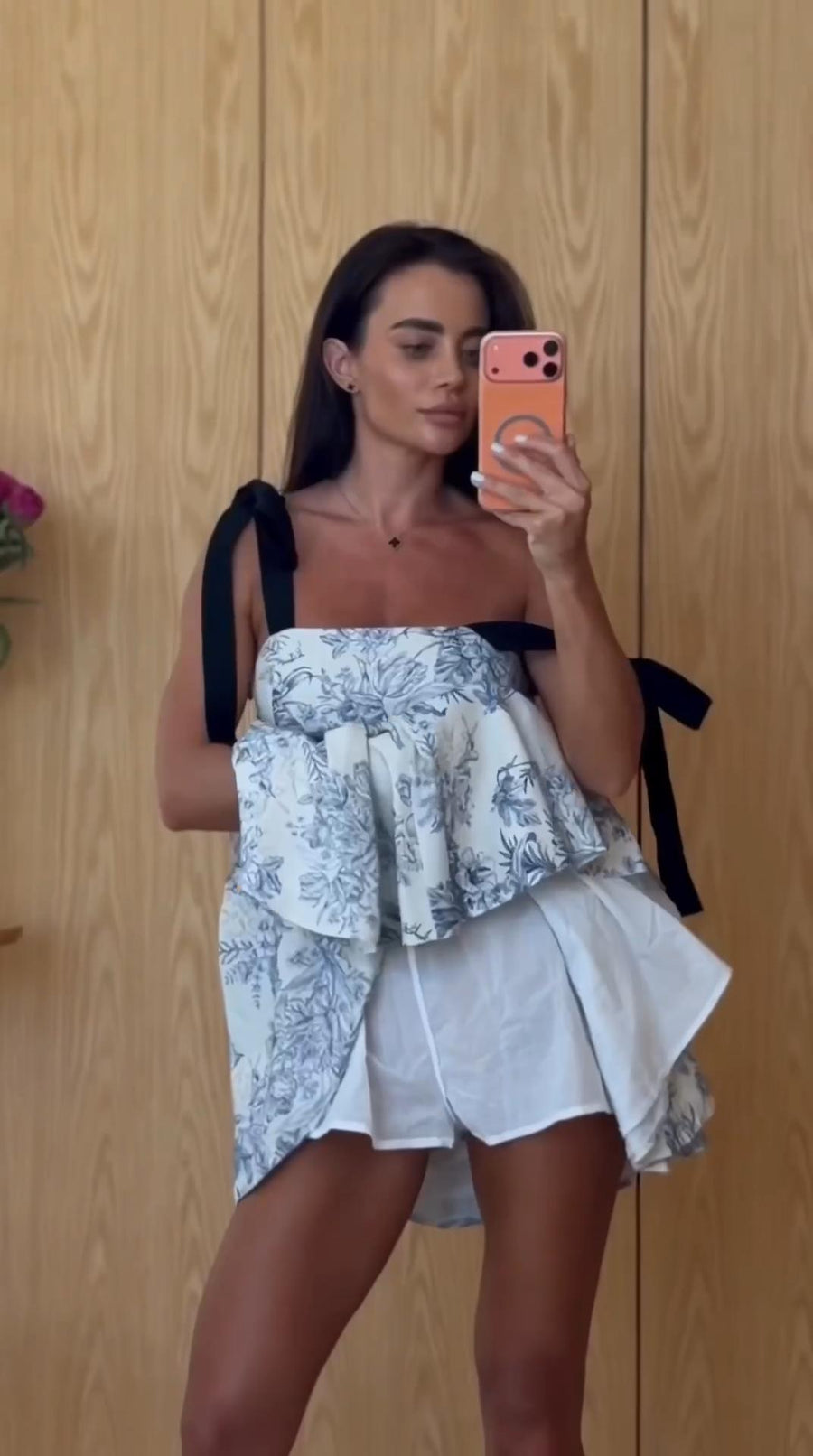 A woman takes a mirror selfie wearing a white ruffled top with a blue floral pattern and black shoulder ties, paired with white shorts, against a light wood-paneled wall.