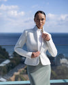 Woman in a white blazer and light gray skirt standing outdoors with a scenic background