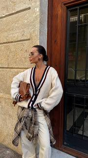 Woman wearing a white cardigan over a plaid shirt with a brown leather bag, standing against a stone wall.