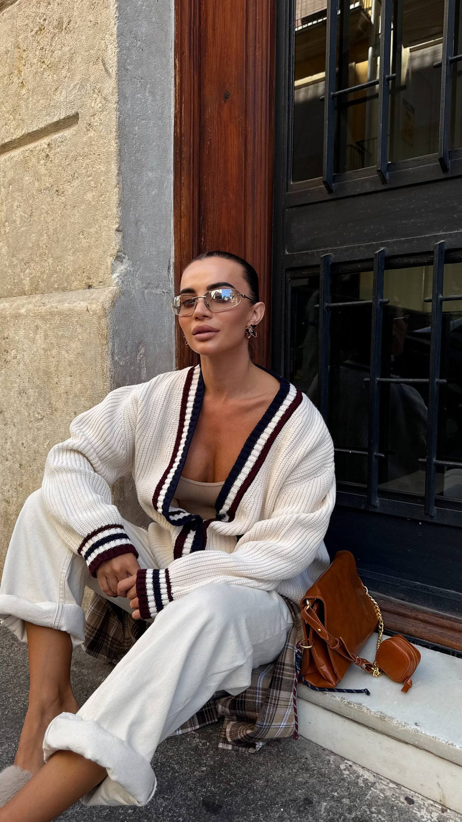 Woman sitting on a step wearing a white cardigan with black stripes, sunglasses, and a brown handbag.