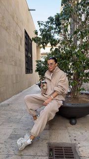 Person in beige outfit sitting on a ledge with a plant and building in the background