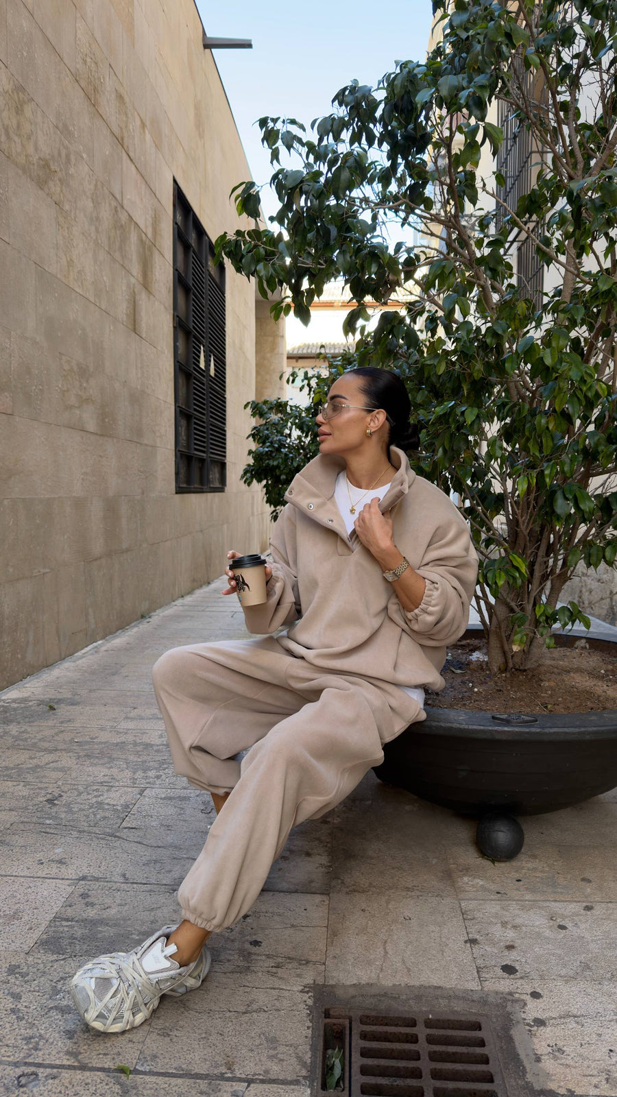 Woman in beige outfit sitting on a city street corner with a coffee cup and tissue.