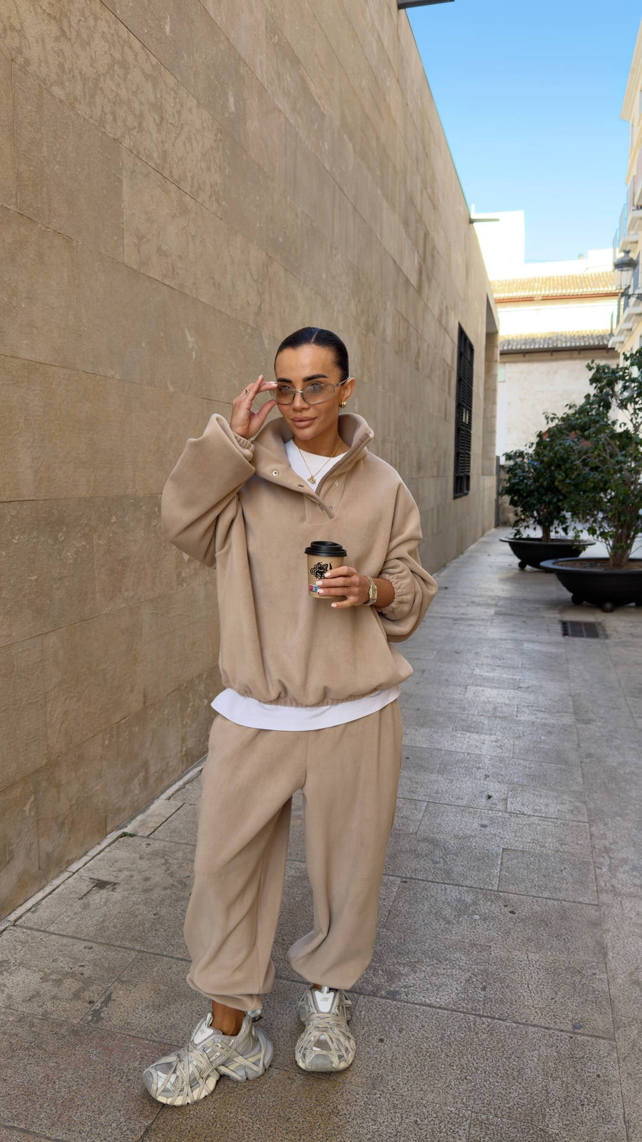 Person wearing a beige tracksuit holding a coffee cup on a sidewalk.