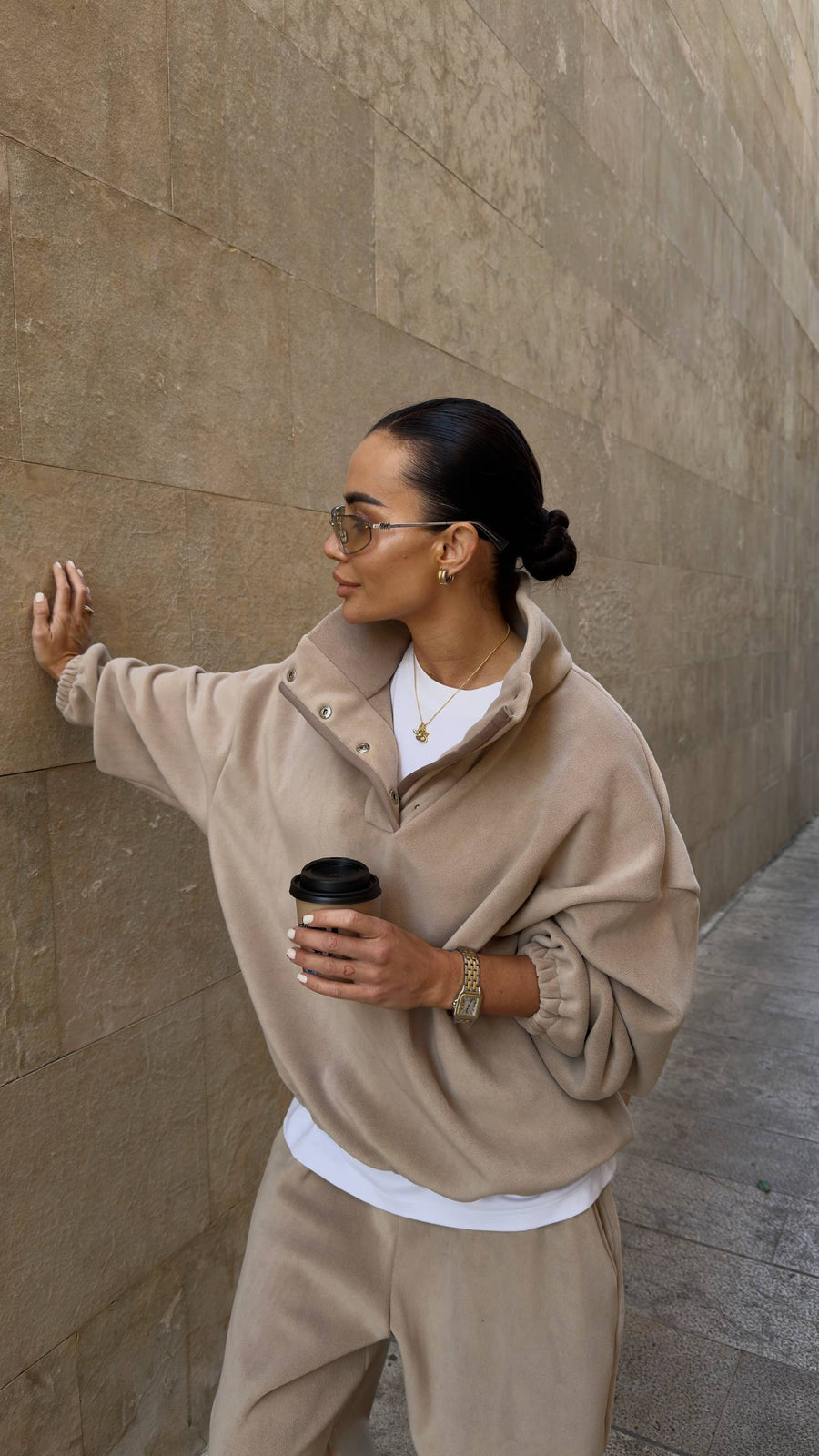 Woman in beige outfit holding a coffee cup against a stone wall.
