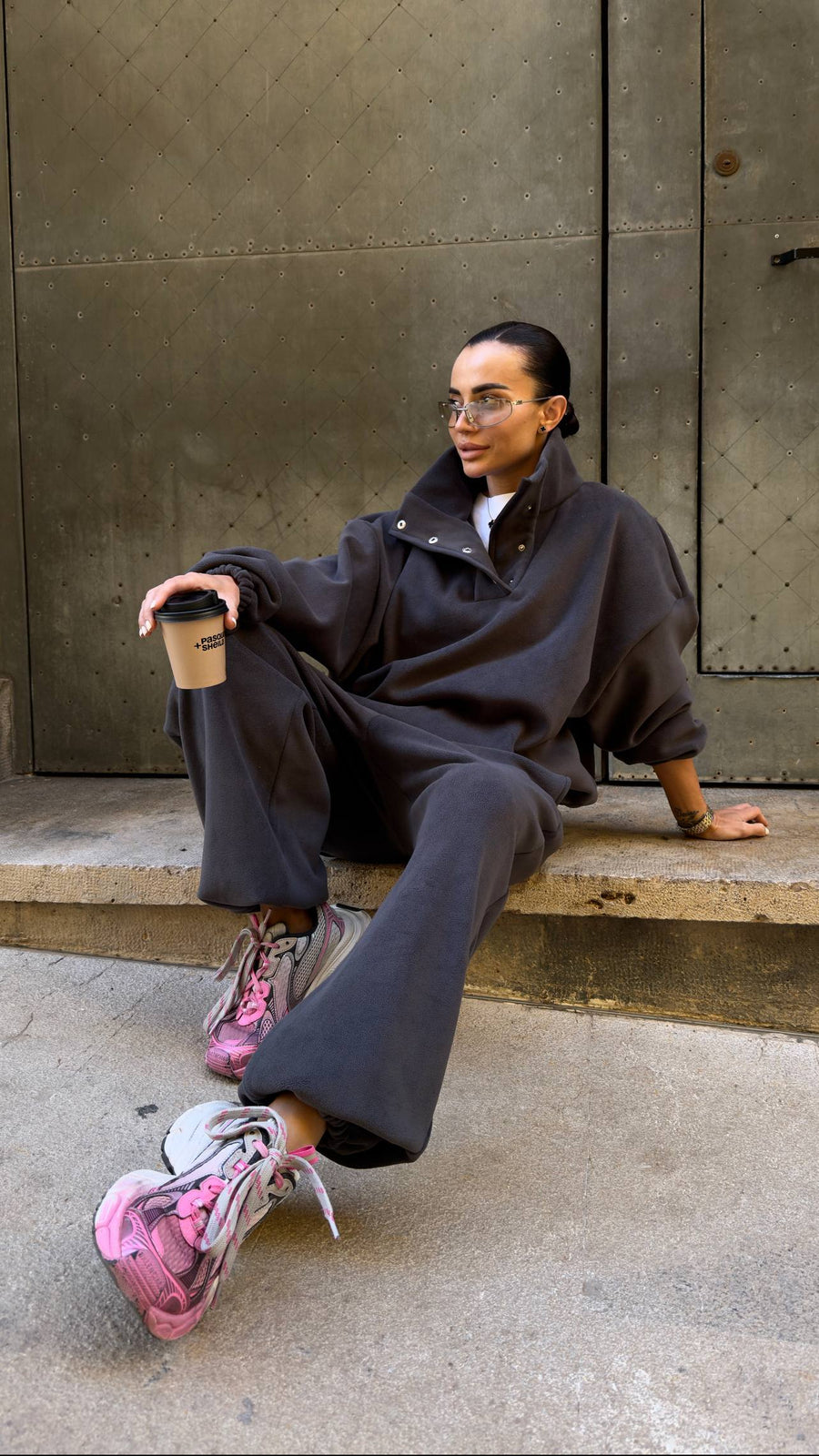 Person sitting on a bench holding a coffee cup, wearing a dark outfit with pink shoes.