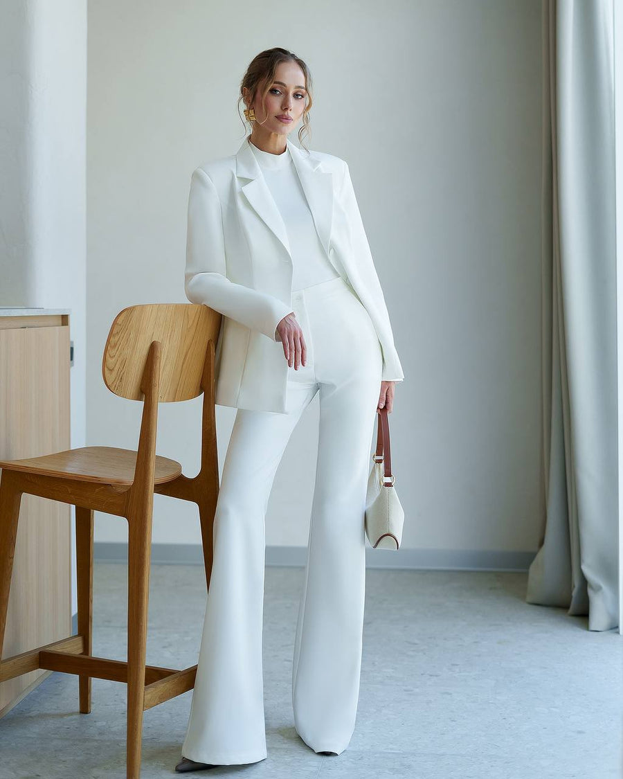 Woman in a white suit standing in a minimalistic room with a wooden chair and light gray walls.