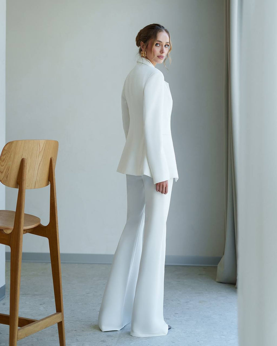 Woman in a white suit standing in a minimalistic room with a wooden chair.