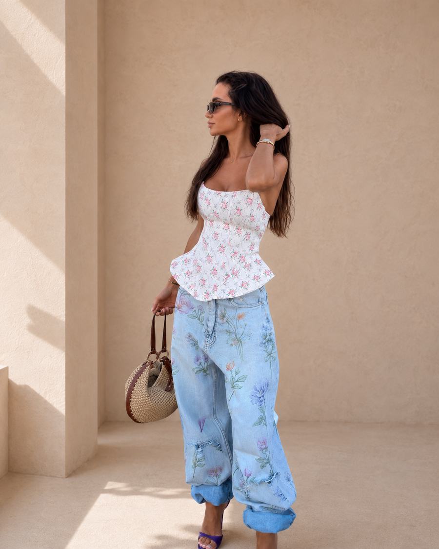 A woman stands in a sunlit beige architectural setting wearing a white strapless top with a pink floral print and light blue wide-leg jeans with a purple floral pattern. She is holding a woven handbag.