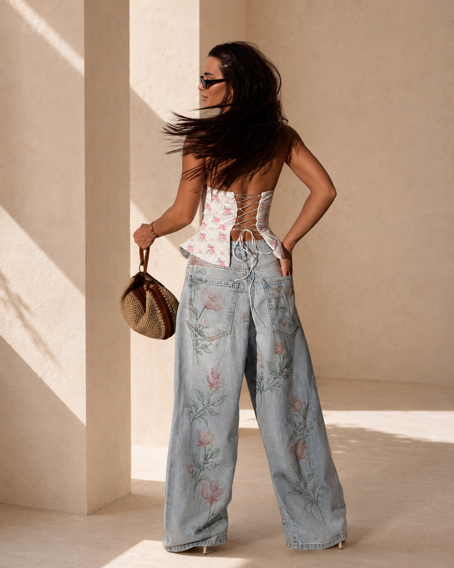 A woman stands in a sunlit architectural setting wearing a floral strapless corset top and wide-leg light-wash denim jeans with pink floral embroidery. She is seen from the back, holding a woven handbag.