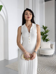 A woman wearing a white sleeveless knit collared dress with a buttoned V-neck, standing in a bright modern interior with white walls, a chair, and plants.