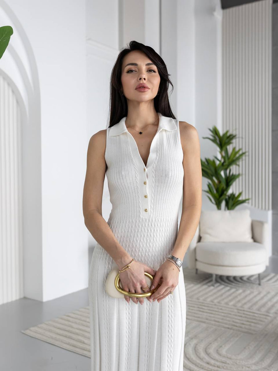 A woman wearing a white sleeveless knit collared dress with a buttoned V-neck, standing in a bright modern interior with white walls, a chair, and plants.