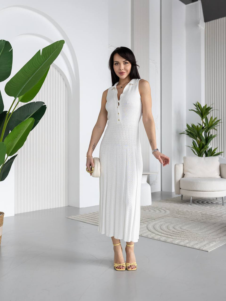 A woman modeling a sleeveless white ribbed midi dress with a V-neckline and front buttons, standing in a bright, modern white interior with plants and a sofa.