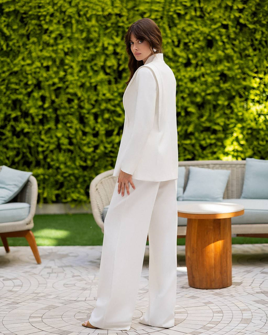 A woman wearing a white two-piece suit with a high collar and wide-leg trousers, standing in a garden patio setting with a green hedge background.
