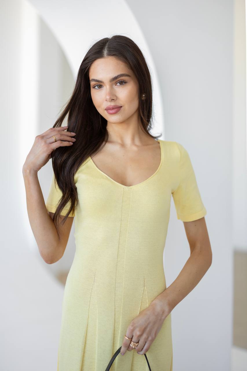 A woman modeling a pale yellow short-sleeve knit dress with a scoop neckline, standing against a bright, minimalist white background.