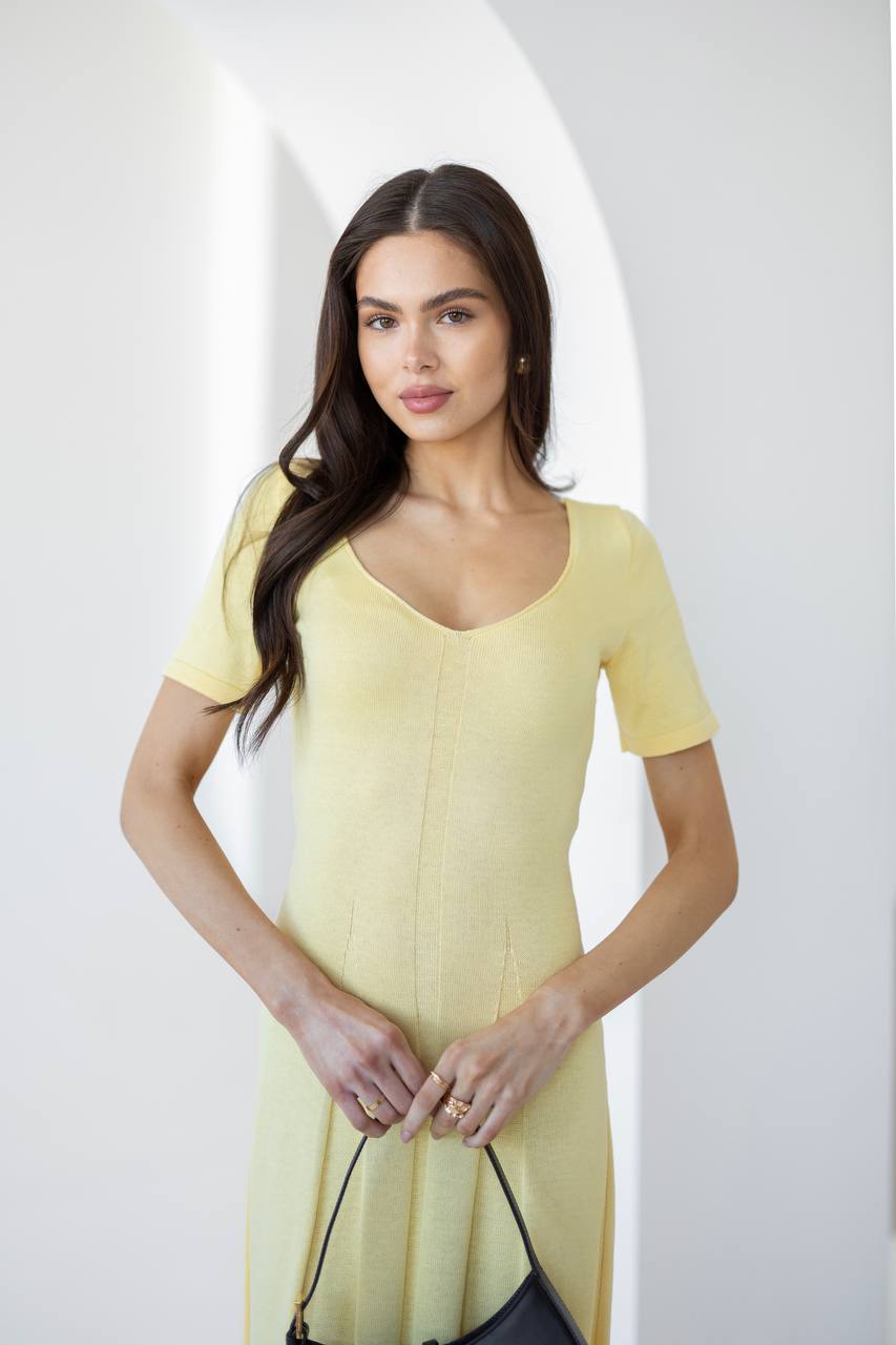 A woman modeling a pale yellow short-sleeve knit dress with a V-neckline, standing against a bright white background with an arched architectural detail.