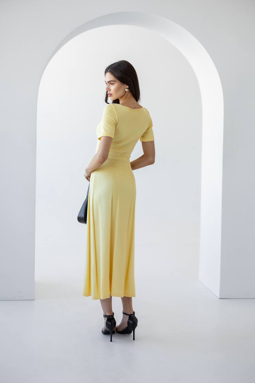 A woman stands in profile wearing a pale yellow short-sleeve midi dress with black high heels, holding a black handbag, set against a minimalist white studio with an arched backdrop.