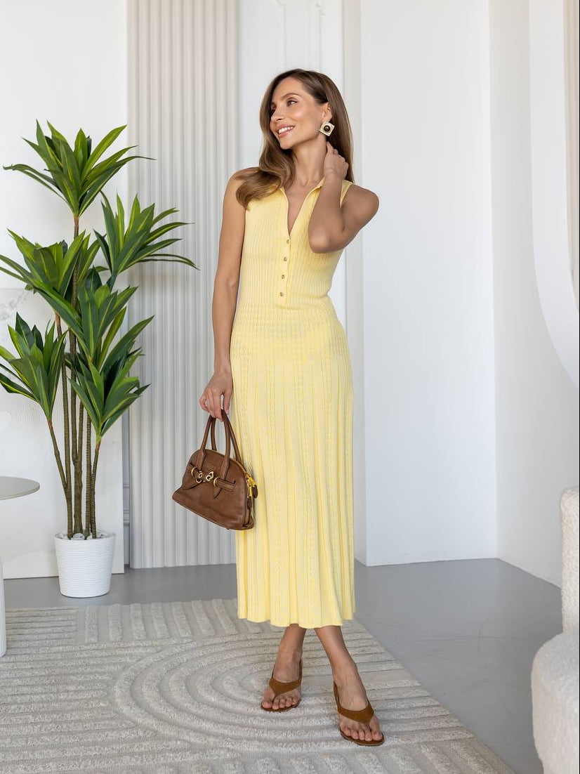 A woman modeling a pale yellow ribbed knit midi dress with a halter neckline and front buttons, standing in a bright, modern room with a potted plant and white columns.